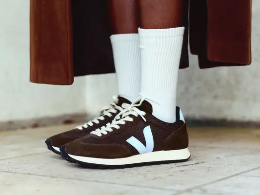 Close-up of person wearing chocolate brown suede sneakers, white crew socks, and brown long coat