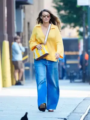 Blake Lively wears a yellow collared top, wide leg jeans, yellow and white crossbody bag, and sunglasses
