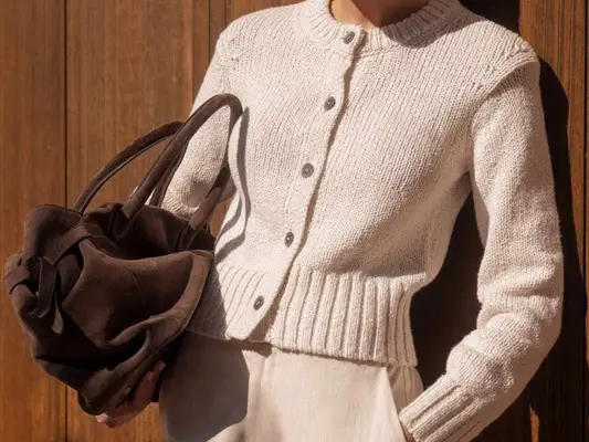 Close-up of model wearing cream sweater and holding chocolate brown suede handbag