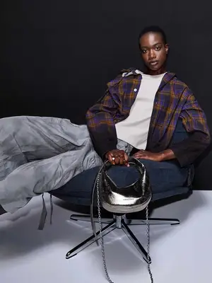 A dark-skinned model is sitting in a navy chair wearing a blue and brown flannel, a white t-shirt, and gray pants. They are holding a silver metallic bag