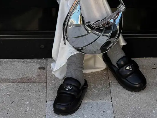 Close-up of person wearing silver handbag, white. maxi skirt, gray ribbed socks, and black Prada loafers