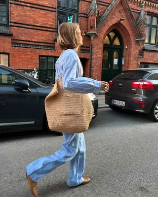 Woman walking wearing a striped button-down, jeans, and a raffia tote bag