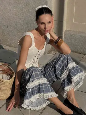 Woman wears white corseted top, blue plaid ruffled pants, black ballet flats, woven handbag, bangles, and white scrunchie