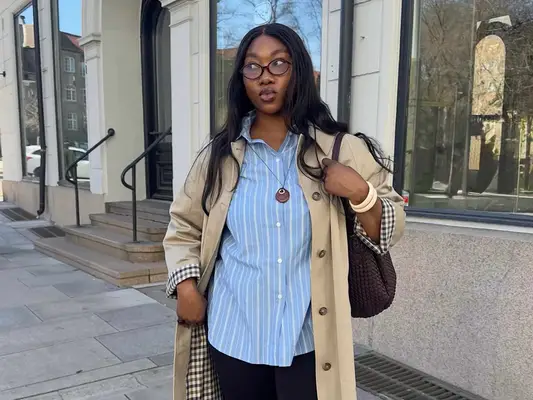 Nnenna Echem wears a striped button-down, trench coat, pendant necklace, woven handbag, and eyeglasses