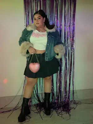Priscilla Del Castillo wearing a denim jacket with fur trim, a Bratz tee, and a black denim pleated skirt while holding a pink heart-shaped purse 