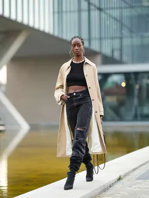 Woman wears gold hoop earrings, a gold chain necklace, a black halter-neck / cropped t-shirt, a beige long coat, a dark brown shiny leather small Telfar handbag, high-waist black denim ripped jeans, and black nylon pointed-toe ankle boots