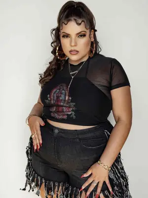 Elle Baez wears a sheer black shirt, graphic tank top, black frayed denim shorts, and oversized hoops 