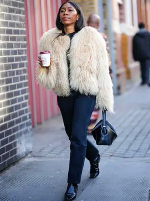 Woman wears a black t-shirt, a beige fluffy sheep jacket, a black shiny leather handbag, black jeans, black leather ankle boots and handbag