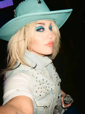Miley Cyrus wears a blue cowboy hat, bedazzled denim shirt, and blue eyeshadow