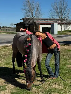 Bella Hadid wears a pink button-down shirt, black crop top, and jeans while posing with a horse