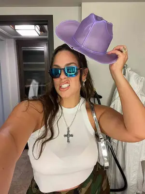 Ashley Graham wears a white ribbed tank top, silver shoulder bag, cross necklace, sunglasses, and purple cowboy hat