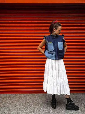 Ella Purnell wearing a denim vest, a white tiered maxi skirt, and black boots. 