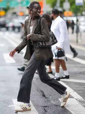 Woman wears blazer, white top, gray jeans, retro Adidas sneakers, and tweed and black handbag