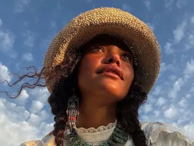 A woman wearing a raffia hat looks up with a serene expression, the sky in the background