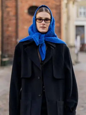 Woman wearing blue knit bandana, sunglasses, and black utilitarian coat