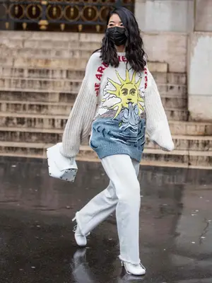 Woman wearing graphic sweater, white jeans, white heels, and handbag