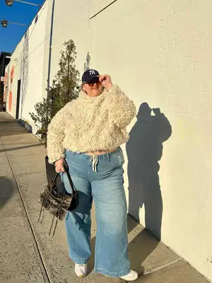 Abby Bible wears a cream-colored textured sweater, light wash jeans, white sneakers, multi-pocketed bag, Yankees cap, and sunglasses