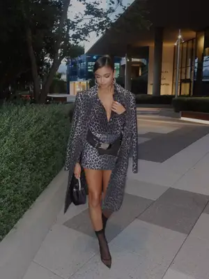 Madison Bailey wears a leopard print mini dress, belt, coat, black handbag, and sheer pointed-toe boots