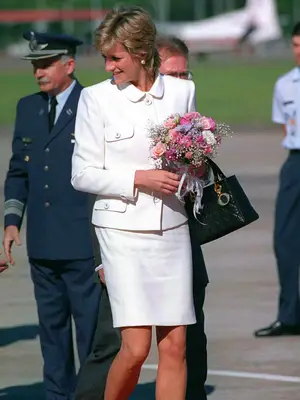Princess Diana walking outside wearing white skirt suit holding pink flowers and black Lady Dior bag