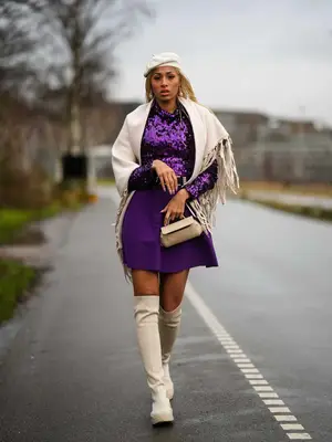 Woman wears a purple sequined top, mini skirt, white fringed jacket, mini handbag, knee-high suede boots, and beret