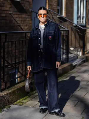 Woman wearing denim jacket, dark wash jeans, gray sweater, white t-shirt, loafers, gold jewelry, and aviator sunglasses