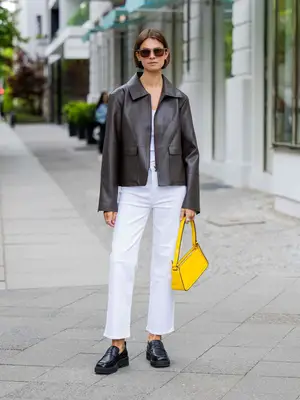 Woman wearing leather jacket, white top, white jeans, yellow handbag, sunglasses, and black loafers