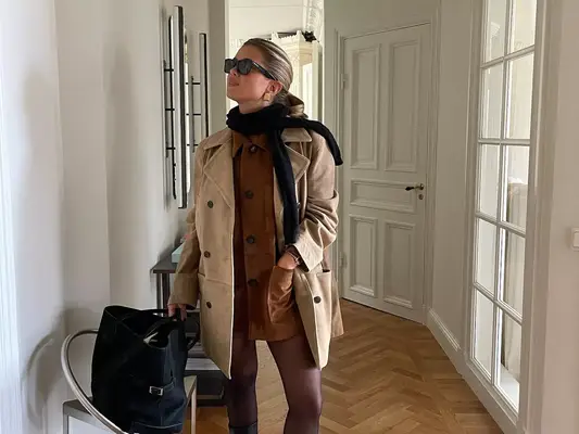 Matilda Djerf wears a brown dress, tights, tan coat, black scarf, sunglasses, and bag
