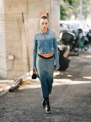 Woman wears a denim cropped jacket and midi skirt with black boots and handbag