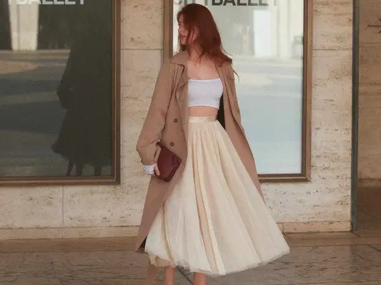 Woman wearing white crop top, beige tulle midi skirt, trench coat, and brown clutch handbag