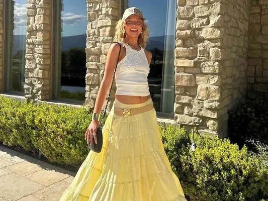 Woman wearing white tank top, trucker hat, butter yellow tiered maxi skirt, and black handbag