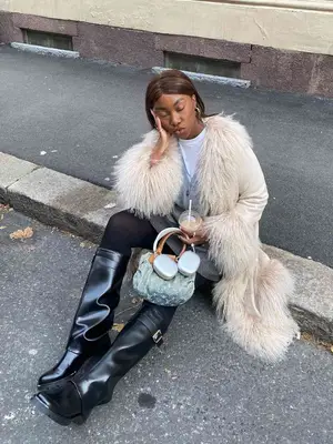 Nnenna Echem wears a cream-colored coat with faux fur trim, bray sweater, white t-shirt, black tights, denim Louis Vuitton bag, black knee-high boots, and silver AirPods Max