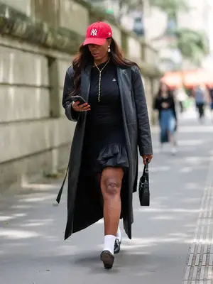 A woman wears New York Yankees red baseball cap hat, white earrings, silver necklace, shiny black leather trench coat, black t-shirt, black mini fluffy skirt, gold rings, black bag, white socks, shiny black loafer leather shoes, 