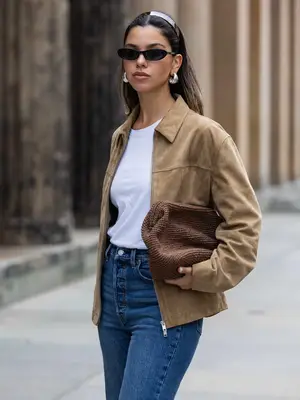 Woman wears cat eye black sunglasses, silver hoops, white tee shirt, brown suede jacket, and blue jeans