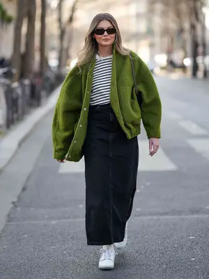 Woman wears black sunglasses, a white and black striped print pattern t-shirt, a green fluffy oversized jacket, a black long denim skirt, white socks, white shiny leather sneakers, a brown shiny leather shoulder bag