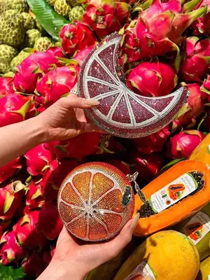 Two hands holding fruit-inspired, bedazzled Judith Leiber handbags in front of a fruit stand