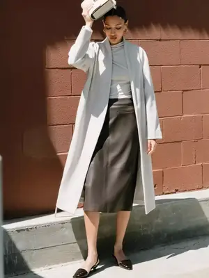 Woman wearing longline coat, brown leather mini skirt, white top, structured handbag, and pointed-toe kitten heels