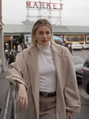 Woman wearing white turtleneck top, beige coat, checkered bottoms, and silver earrings