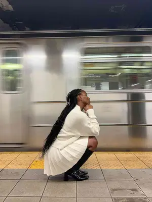 Woman crouching wearing black square-toe boots, white sweater and skirt, and claw clip on subway platform