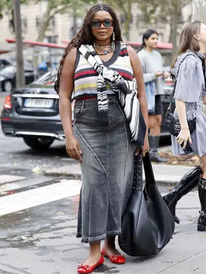 Gabriella Karefa Johnson wears a colorful striped sweater vest, black and white striped tied cardigan, denim maxi skirt, red flats, black hobo bag, and oversized square sunglasses