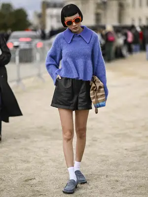 Kiwi Lee wears layered blue sweaters, black leather shorts, striped tote, gray moccasins, and red sunglasses