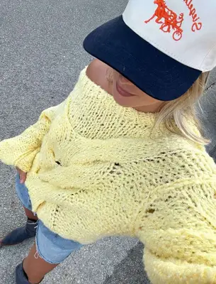 Woman wearing bright yellow open-knit sweater, denim shorts, cowboy boots, and baseball cap