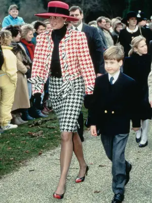 Princess Diana walking outside with Prince William wearing red and black brimmed hat with black caging, red printed blazer over black top, black and white printed skirt, and red and black heels