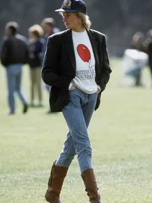 Princess Diana walking outside on grass wearing navy blue hat, black blazer, white sweater with red balloon print that says london foundation, blue jeans, and brown boots