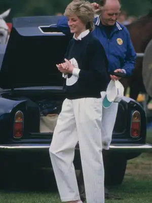 Princess Diana standing by trunk of car smiling wearing white striped slacks, black sweater with white button up underneath