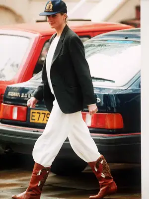 Princess Diana wearing a blue ball cap, black blazer, white t-shirt, and white flowing pants tucked into brown cowboy boots