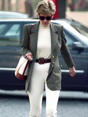 Princess Diana walking outside wearing sunglasses and purse over shoulder, with green blazer, cream turtleneck tucked into white jeans with brown belt