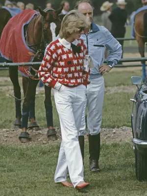Princess Diana wears her iconic black sheep sweater paired with white trousers, red shoes, white collared top with black bow, and sunglasses