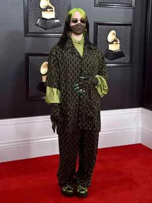 Billie Eilish wears an oversized black and green Gucci logo pants set with fingerless gloves, sneakers, and a sheer mask