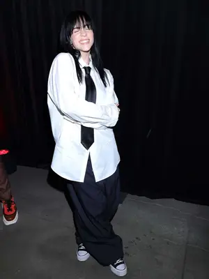 Billie Eilish wears an oversized white button-down shirt, black tie, and baggy black pants to the 