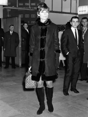 Audrey Hepburn in a black fur-trimmed coat and black boots, carrying a Louis Vuitton Speedy bag